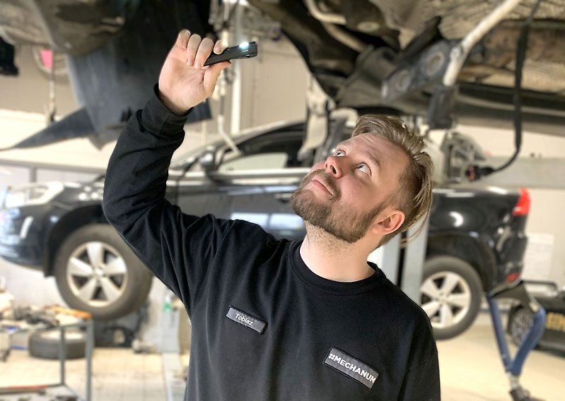 Man står i bilverkstad under ett fordon som han kontrollerar