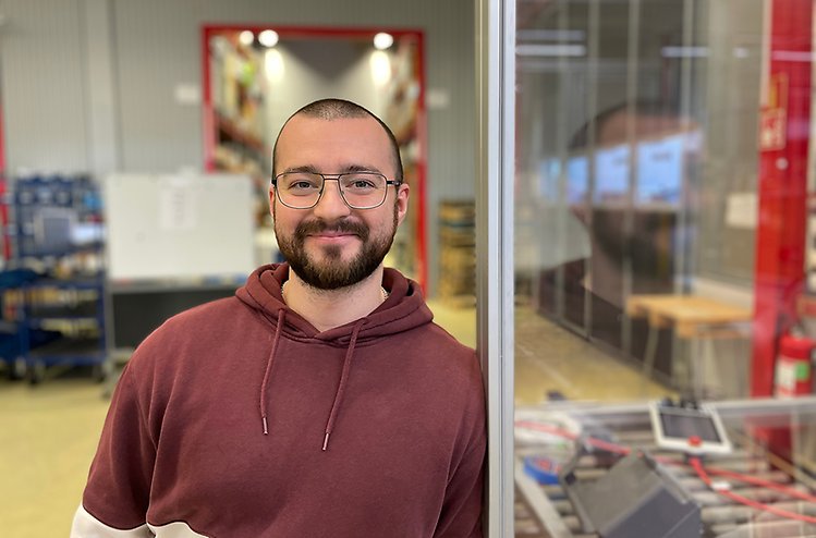 Glad yngre man med mörkt hår, skägg och glasögon står i fabrik och tittar in i kameran.