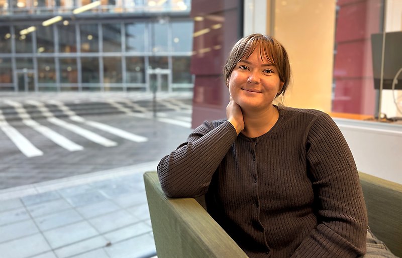 Tjej sitter i soffa och tittar in i kameran. Hon har ett stort fönster bakom sig och intill står det en arbetsplats.