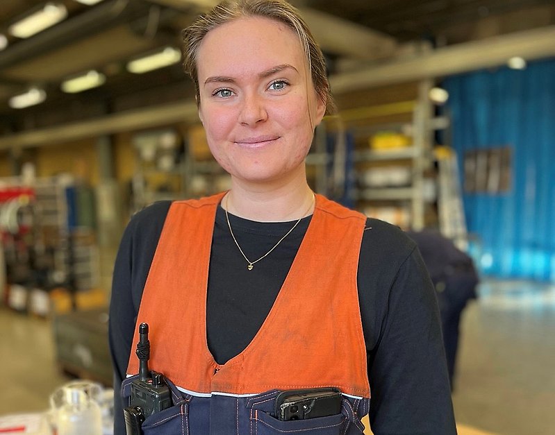 Tjej står i verkstad. Hon är klädd i arbetskläder och tittar in i kameran.